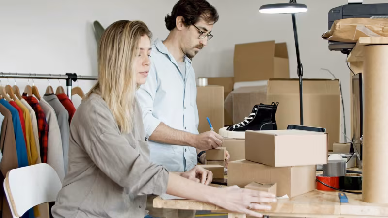 A couple packing shoes and other items into boxes at a table beside a clothes rail