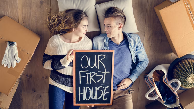 A couple lying on the floor holding a chalkboard reading Our first house, seen from above