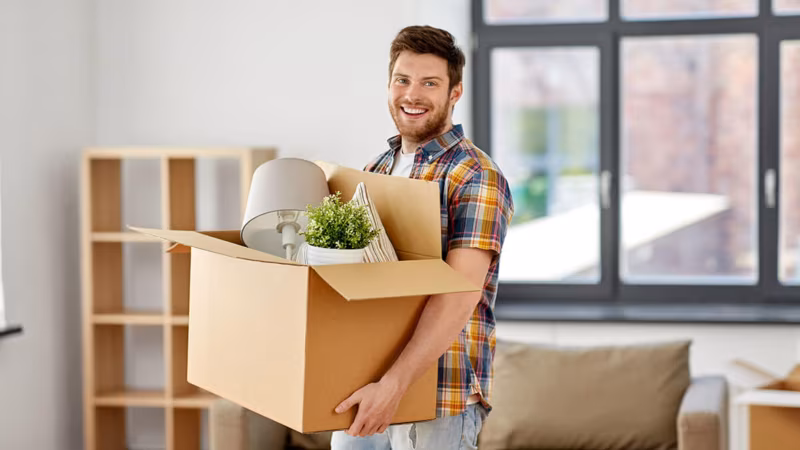 A man carrying an open box of belongings through a bright room