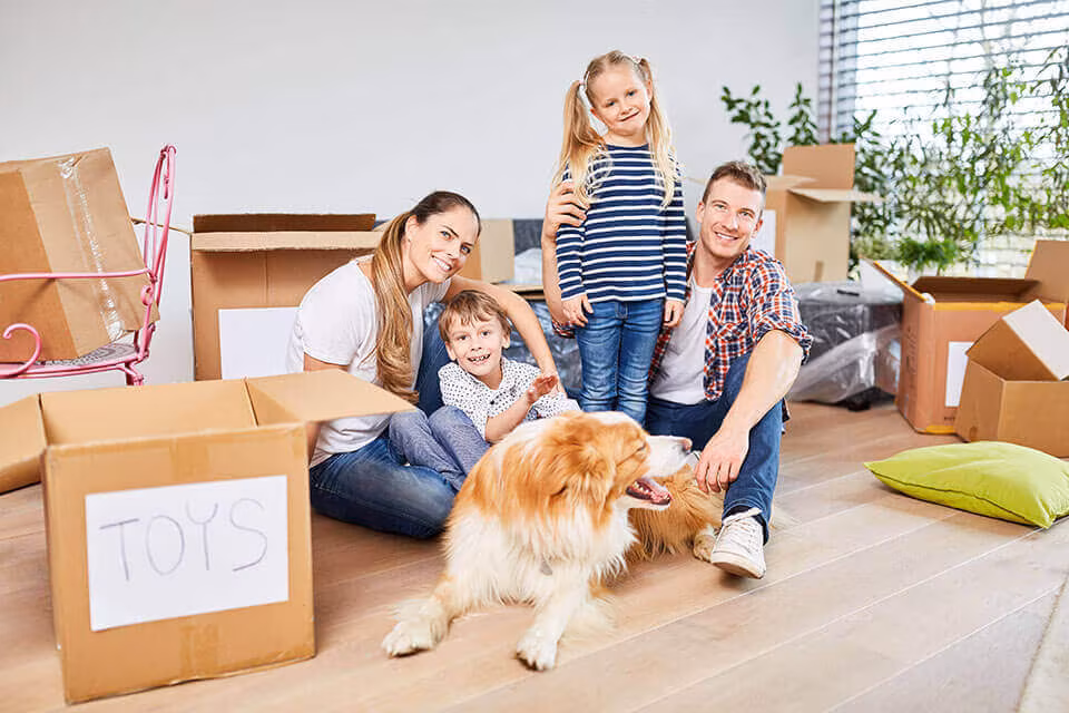A family sitting on the floor with their dog among cardboard boxes, one labeled Toys