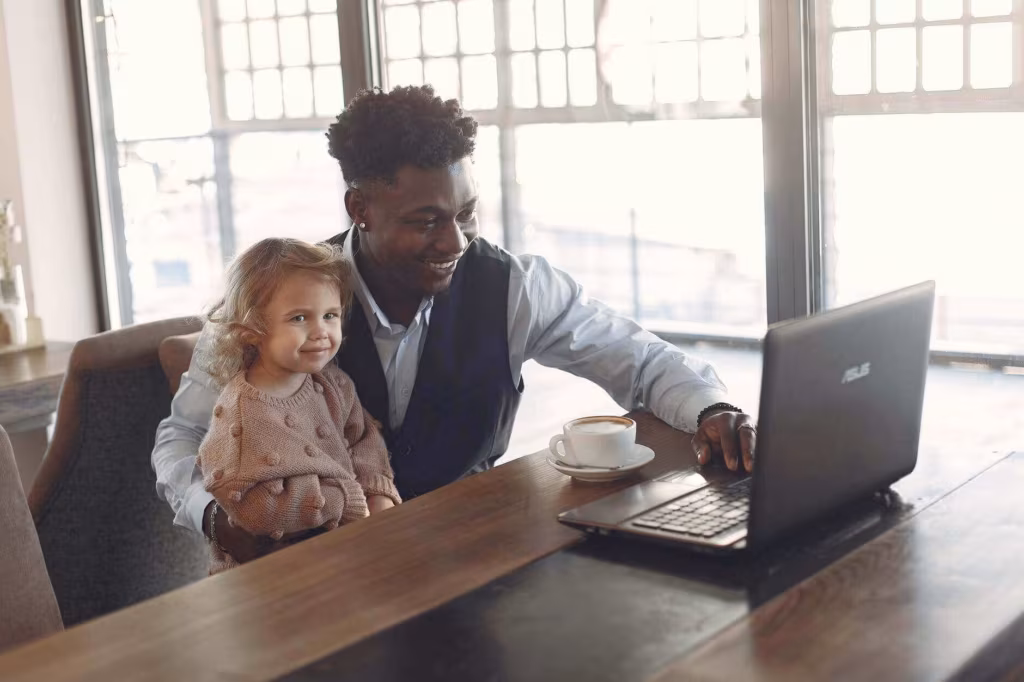 A man working on a laptop at a table with a small child on his lap