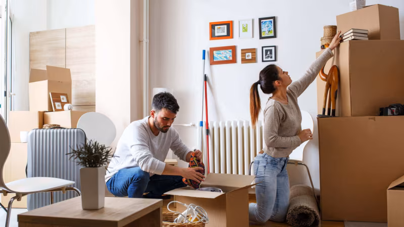 A couple packing up a living room with cardboard boxes and a rolled rug