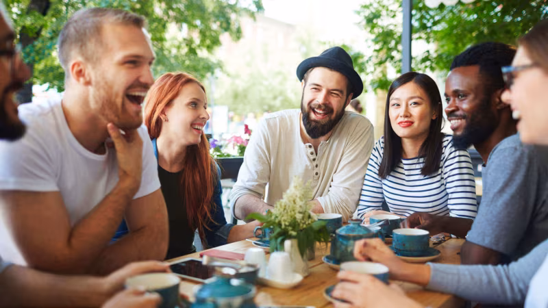 A group of friends laughing around an outdoor table set with coffee cups