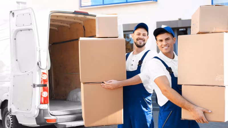 Two movers in blue carrying stacks of cardboard boxes beside a van