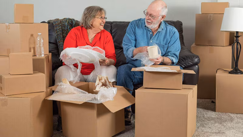 An older couple sitting on a sofa wrapping china, surrounded by cardboard boxes