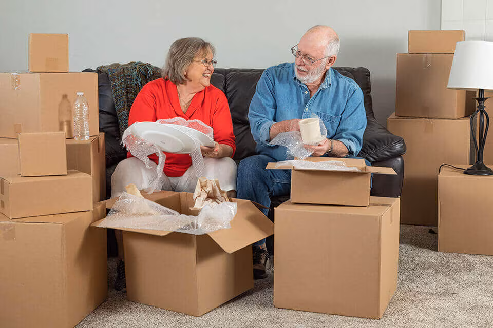 An older couple sitting on a sofa wrapping china, surrounded by cardboard boxes