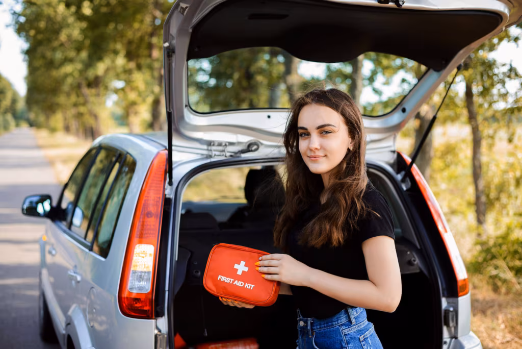 A young woman at the open boot of a car holding a red first-aid kit