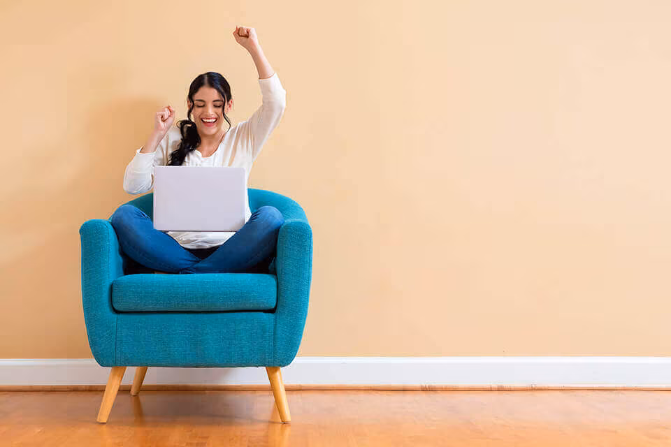 A woman with a laptop in a teal armchair, raising one arm in celebration