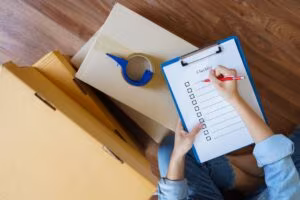 Seen from above, a person filling in a checklist on a clipboard beside a flat cardboard box and a roll of packing tape