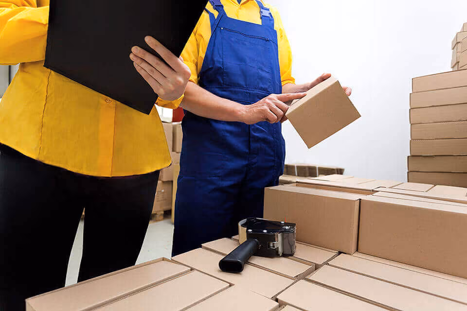 Two people in work clothes checking a clipboard among stacks of flat-packed cardboard boxes