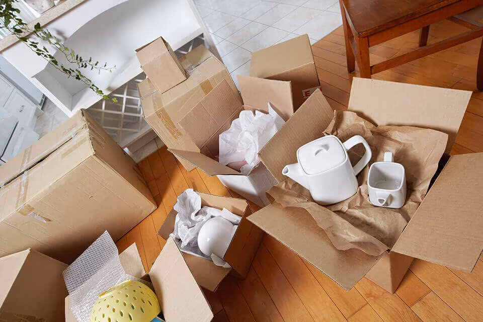Open cardboard boxes on a wooden floor with crockery and a teapot wrapped in paper