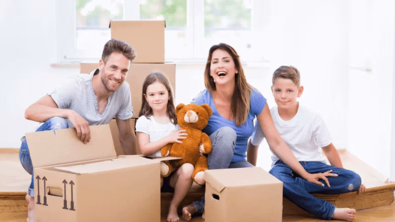 A family of four sitting on the floor unpacking boxes, one child holding a teddy bear