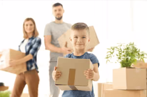 A family carrying cardboard boxes into a new home, with a boy in front holding one