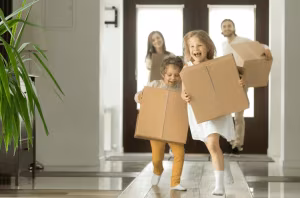 A family walking through a doorway carrying cardboard boxes, the children in front