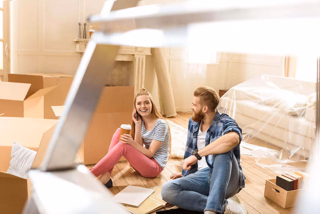 A couple sitting on the floor of a part-packed room with coffee, boxes and furniture under plastic sheeting
