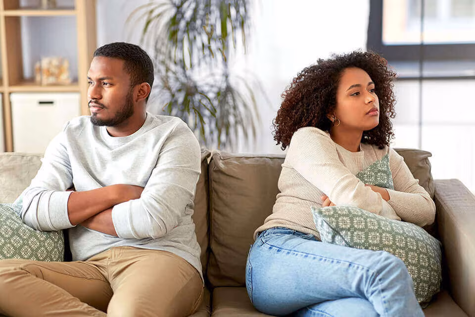 A couple sitting at opposite ends of a sofa with their arms crossed, looking away from each other