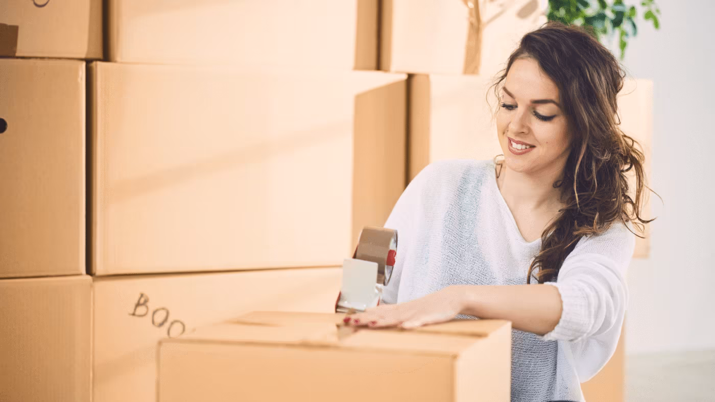 A woman taping a box labeled books among stacked cardboard boxes