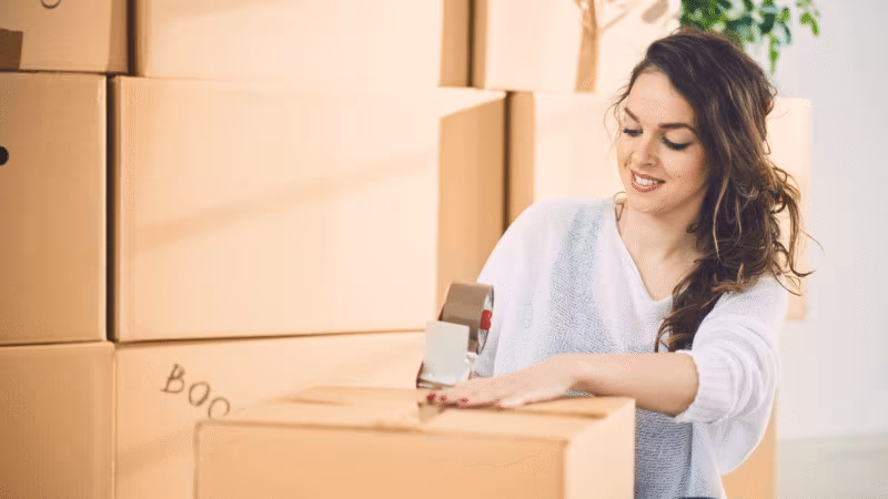 A woman taping a box labeled books among stacked cardboard boxes