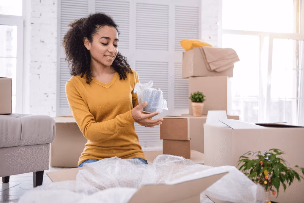 A young woman sitting on a bed wrapping items among cardboard boxes