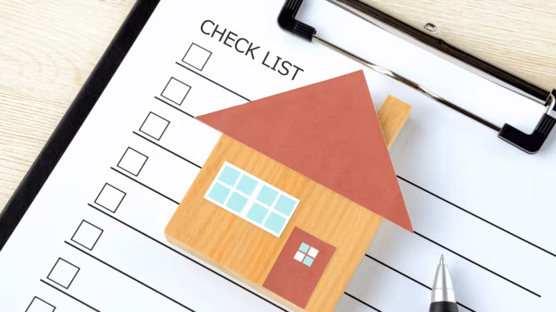 A paper cut-out house on a clipboard checklist with empty tick boxes and a pen