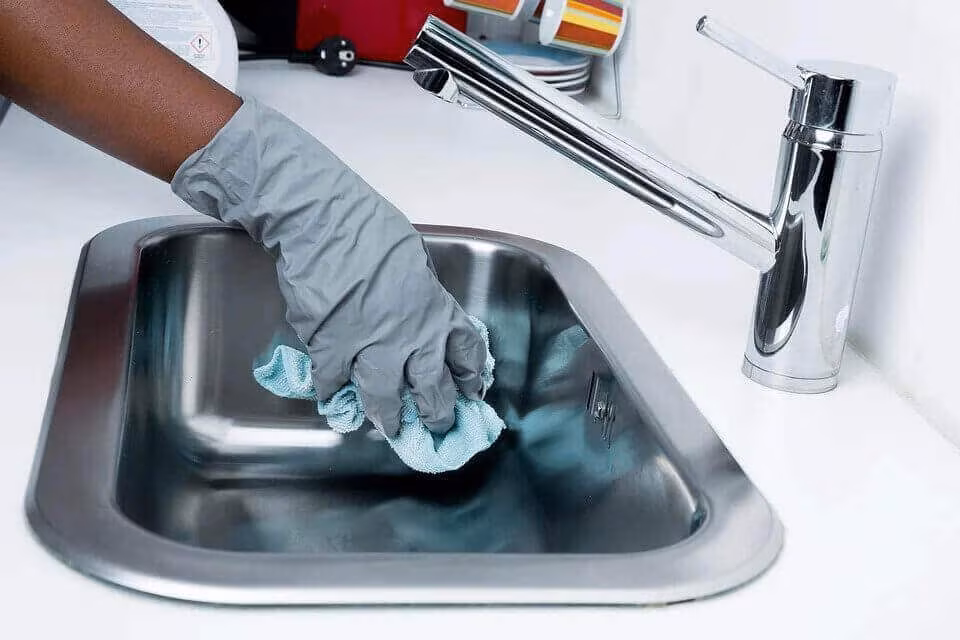 A gloved hand wiping a stainless steel sink with a cloth