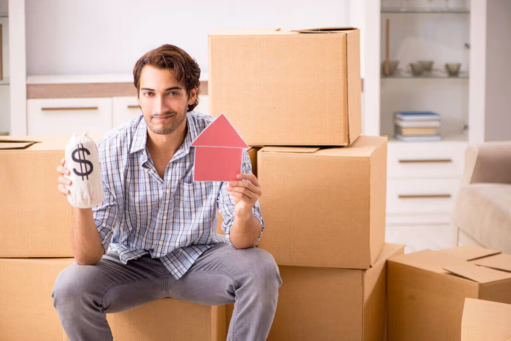 A man sitting on a cardboard box holding a paper dollar sign and a small red paper house