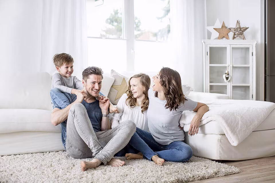 A family of four sitting together on the floor and sofa of a bright living room