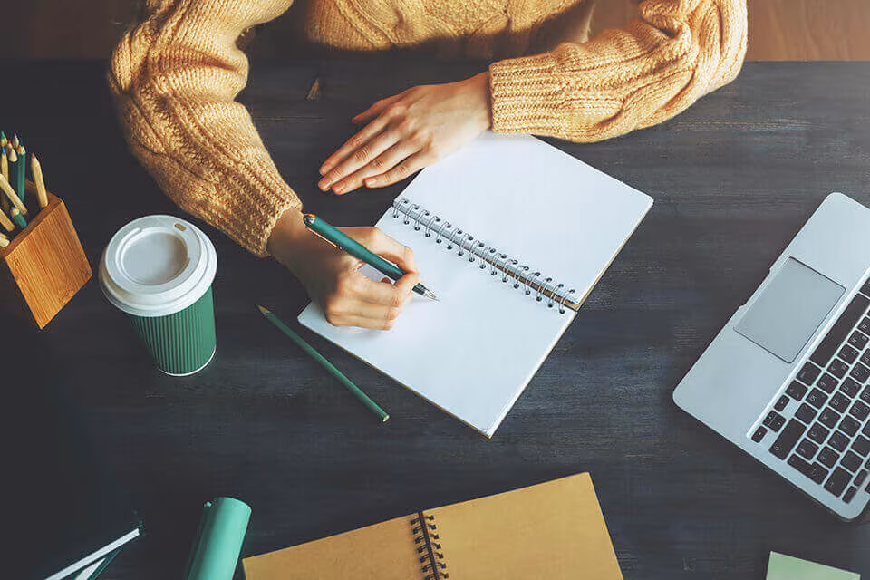 Seen from above, hands writing in a notebook beside a coffee cup and a laptop