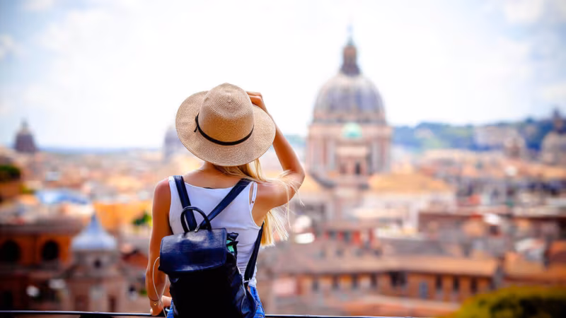 A woman with a backpack and sun hat looking out over a city with a domed cathedral