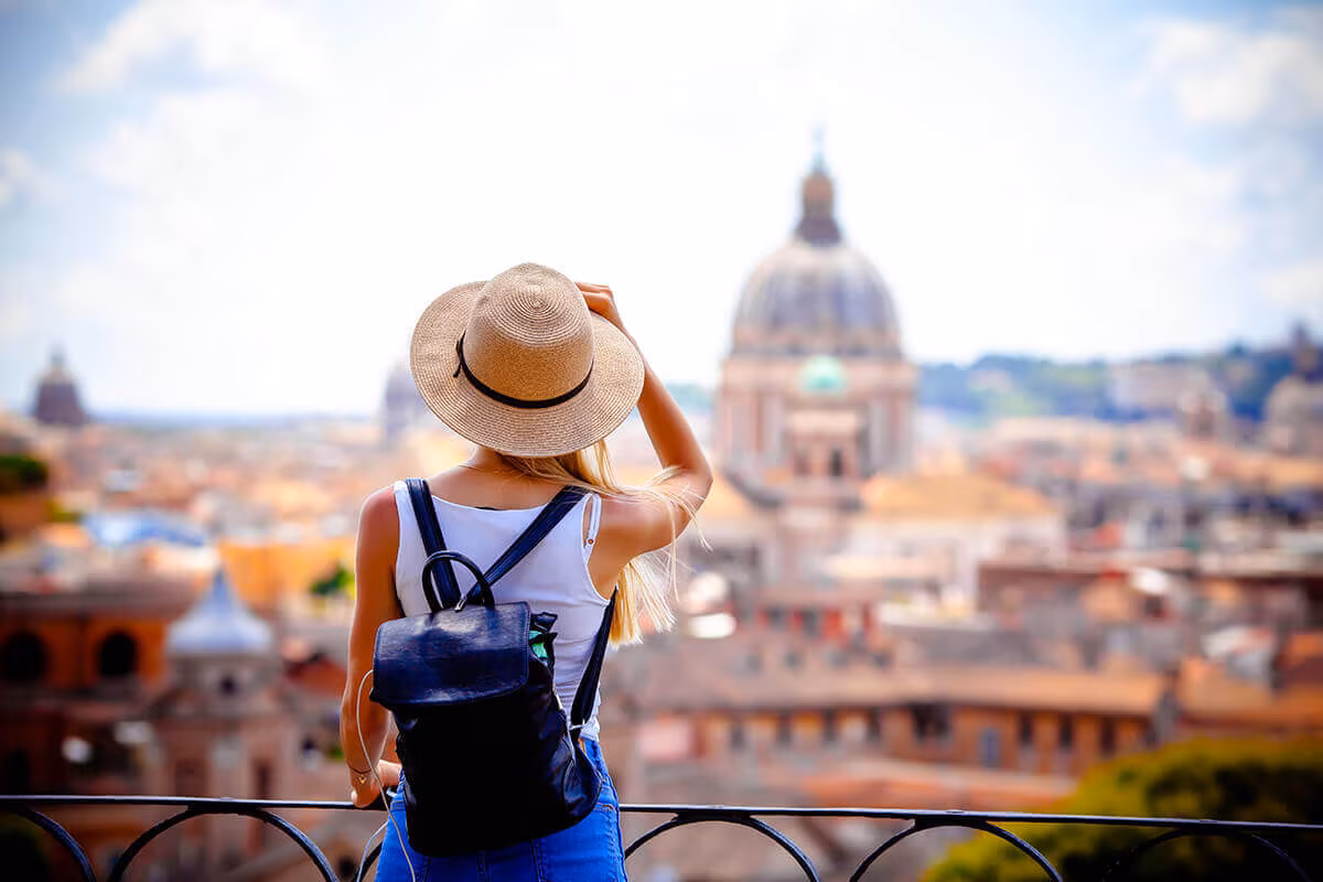 A woman with a backpack and sun hat looking out over a city with a domed cathedral