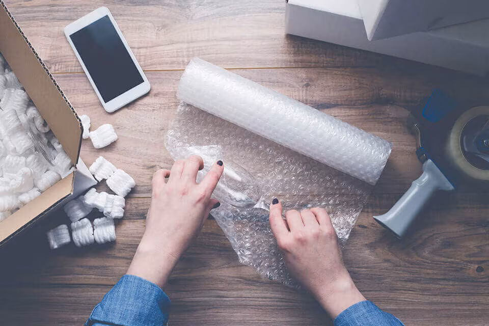 Seen from above, hands wrapping a glass in bubble wrap beside a phone, tape and packing peanuts