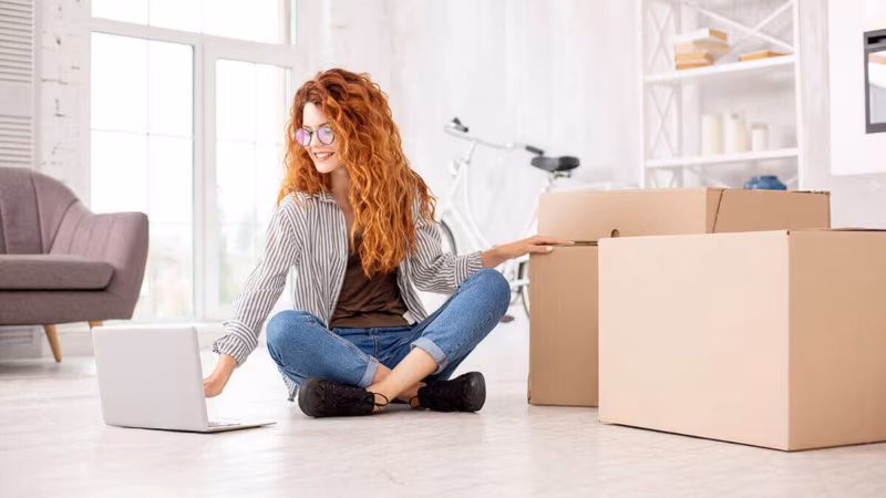 A woman sitting cross-legged on the floor with a laptop beside cardboard boxes