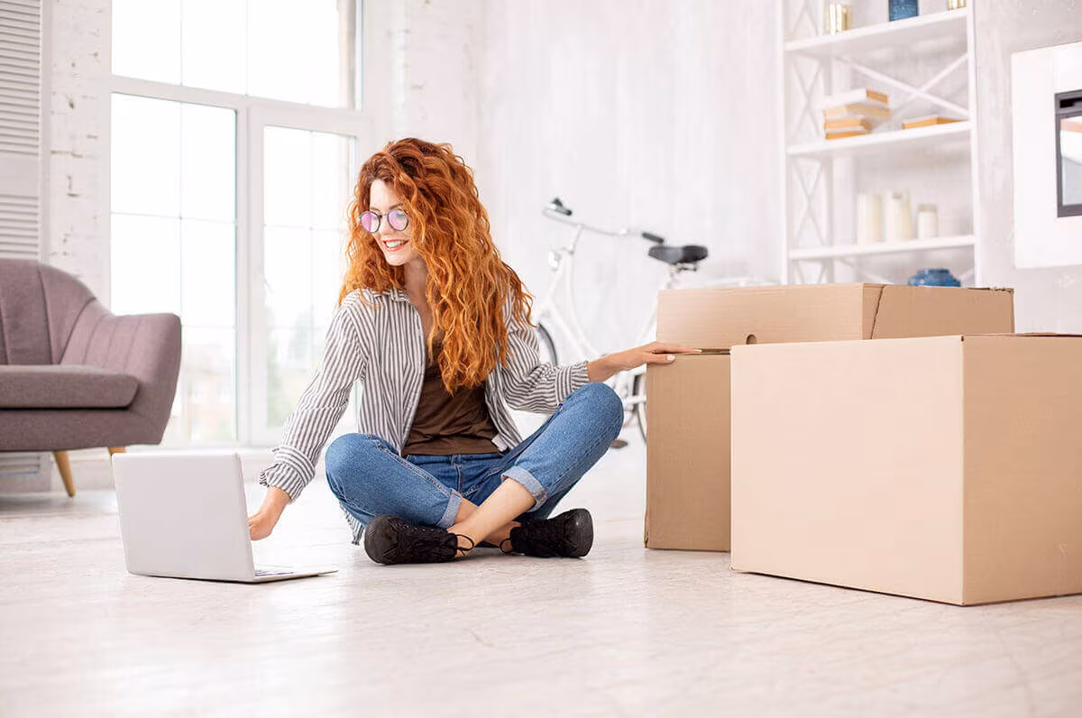 A woman sitting cross-legged on the floor with a laptop beside cardboard boxes