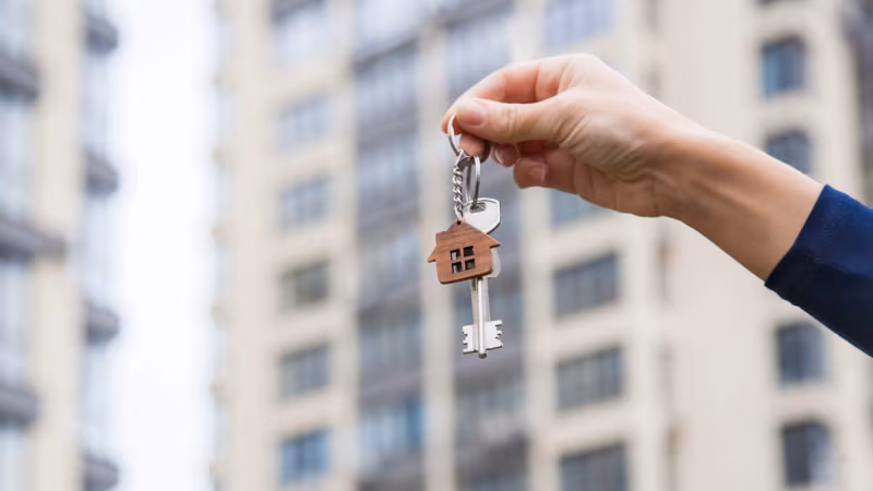 A hand holding keys with a house-shaped fob in front of an apartment block