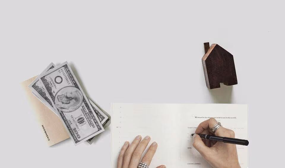 Seen from above, a hand signing a document beside banknotes and a small wooden house