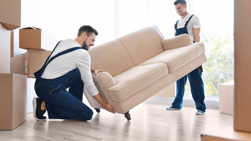 Two movers lifting a beige sofa in a bright room with cardboard boxes