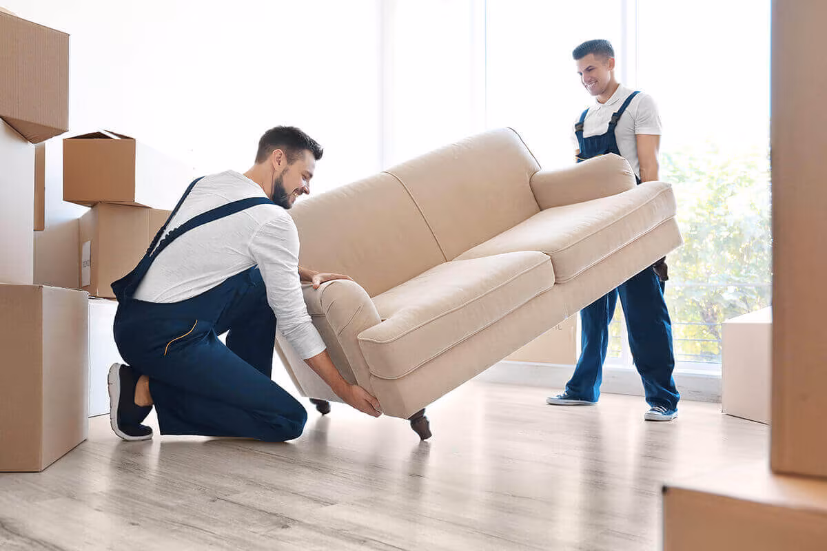 Two movers lifting a beige sofa in a bright room with cardboard boxes