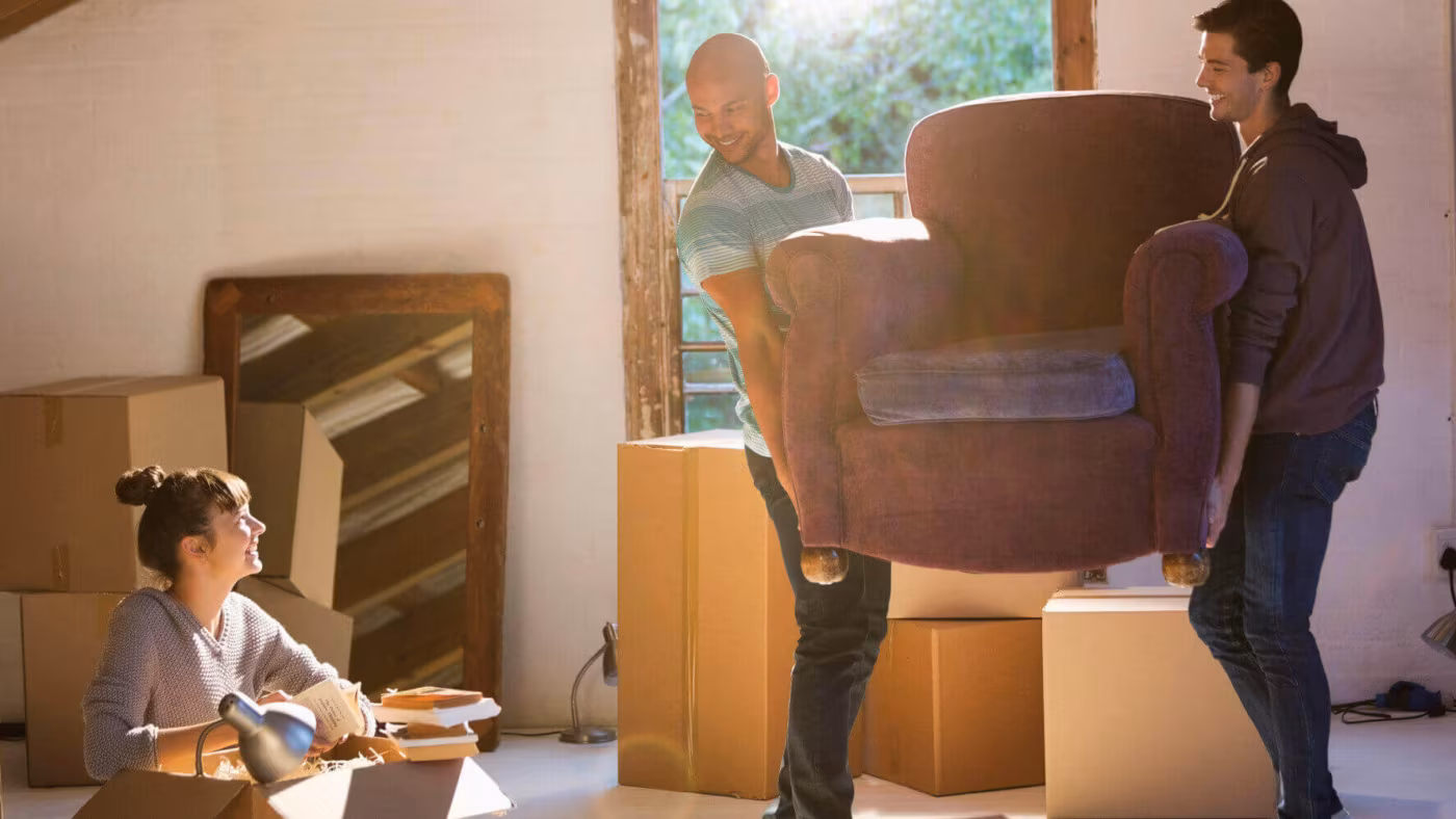 Two people carrying an armchair through a room of boxes while a third packs