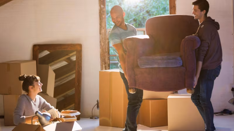 Two people carrying an armchair through a room of boxes while a third packs