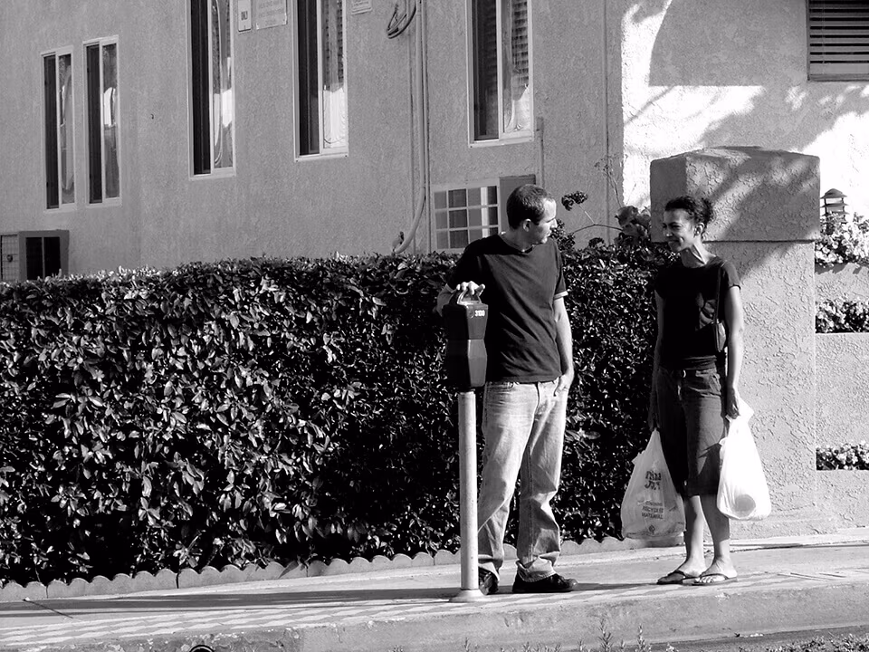 Two people talking on a pavement beside a hedge, in black and white