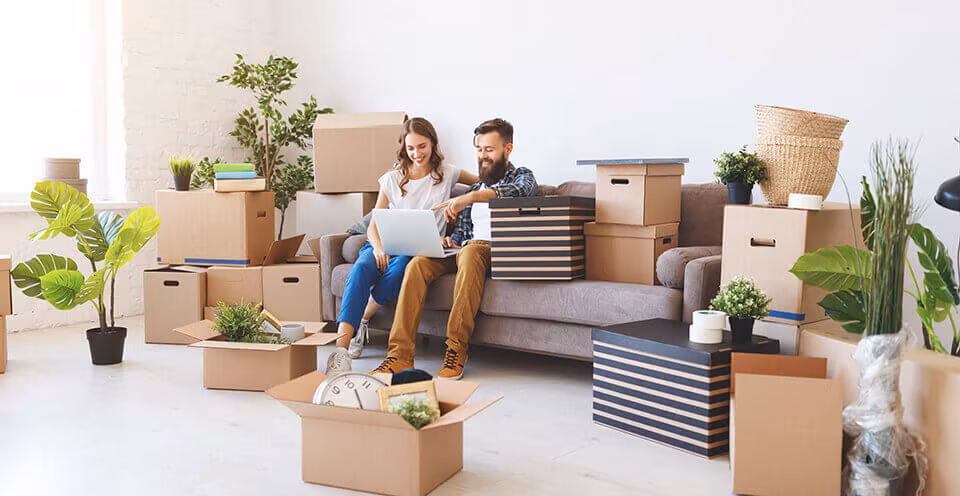 A couple sitting on a sofa with a laptop among cardboard boxes and houseplants