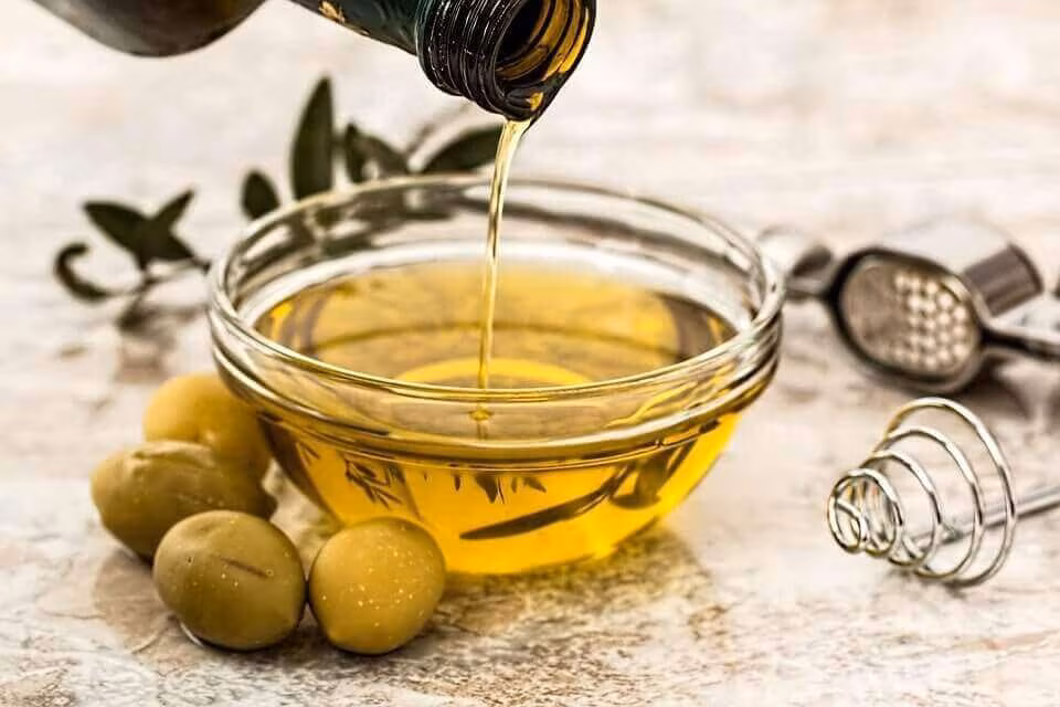 Olive oil being poured into a glass bowl, with olives and a sprig beside it