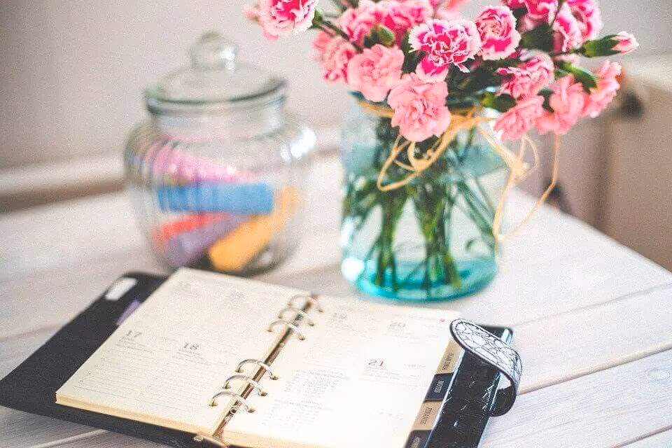 An open ring-binder planner on a desk beside a jar of colored pens and a vase of pink carnations