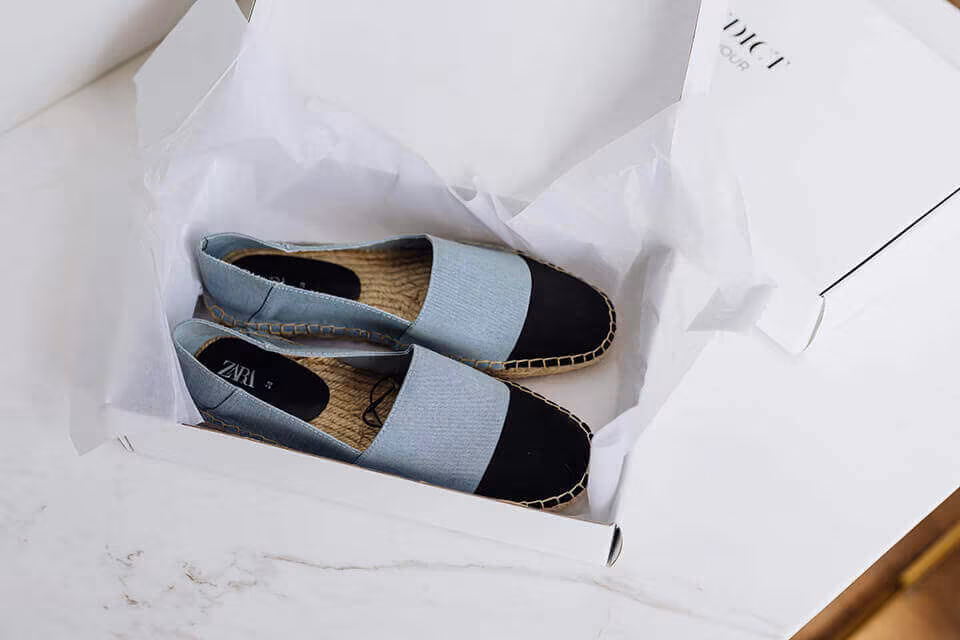 A pair of black espadrilles in an open shoebox lined with tissue paper