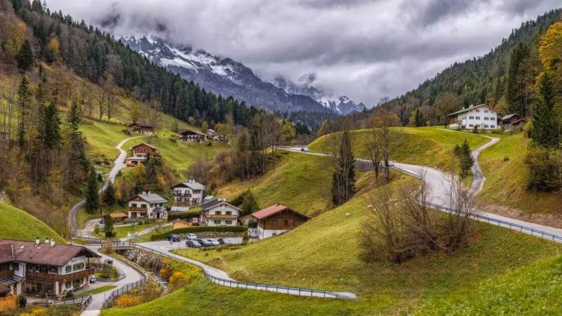A green alpine valley with scattered farmhouses below cloud-covered mountains