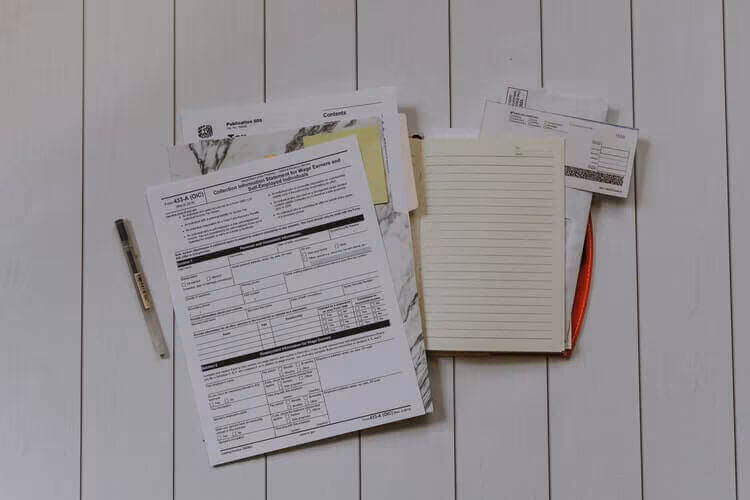 Printed forms, a notepad and a pen laid out on a white wooden surface