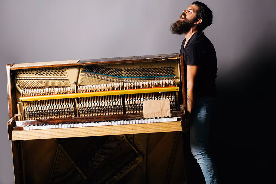 A man leaning on the wall beside an upright piano with its front panel removed and the strings exposed
