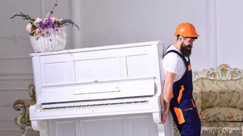 A mover in overalls and a hard hat moving a white upright piano