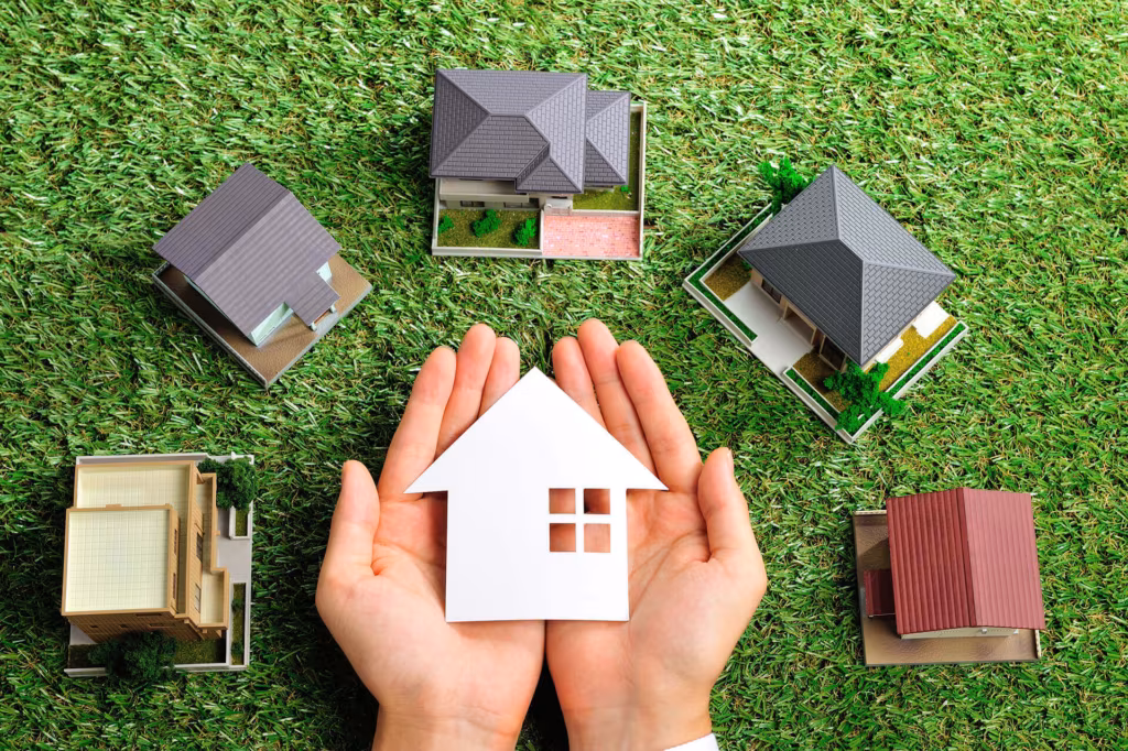 Two hands holding a small white paper house, surrounded by architectural models on grass