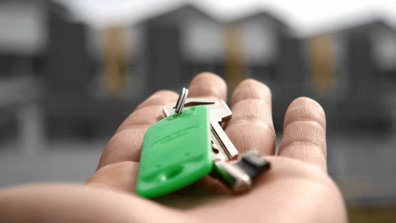 An open palm holding a key with a green fob in front of apartment buildings